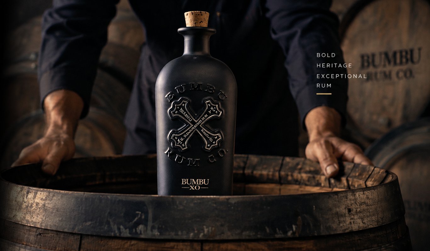 A bottle of Bumbu XO rum resting on an oak barrel in a dimly lit distillery, with a craftsman's hands resting on the barrel in the background.