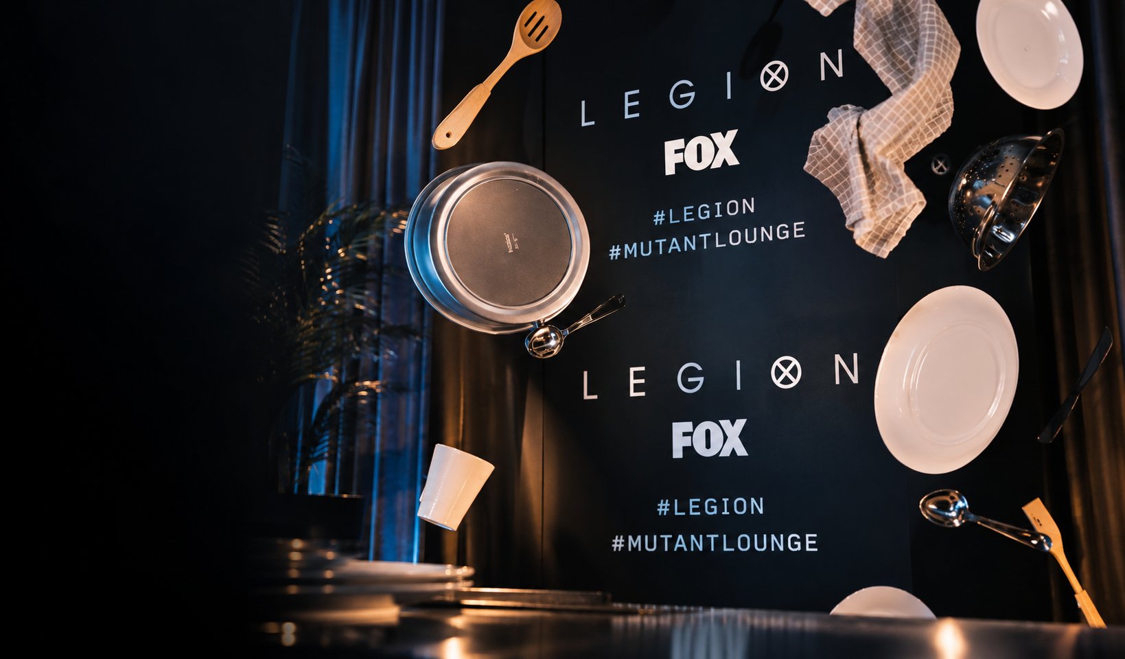 Legion and Fox branding printed on a dark backdrop at the Mutant Lounge launch event, with kitchen props including plates, a saucepan and utensils suspended in front of it.