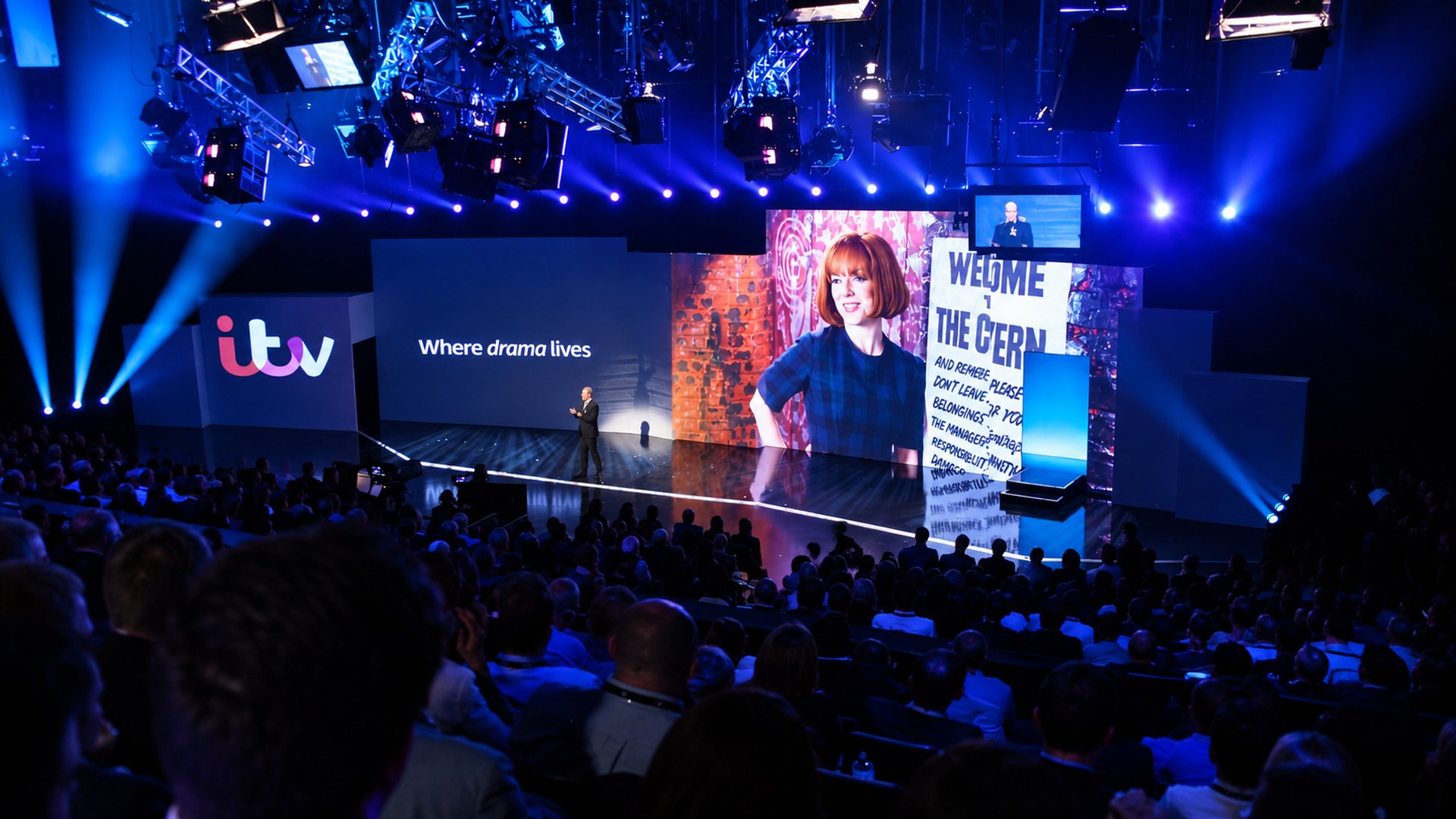 A presenter on the ITV Upfronts stage in front of screens reading Where drama lives, with programme artwork behind.