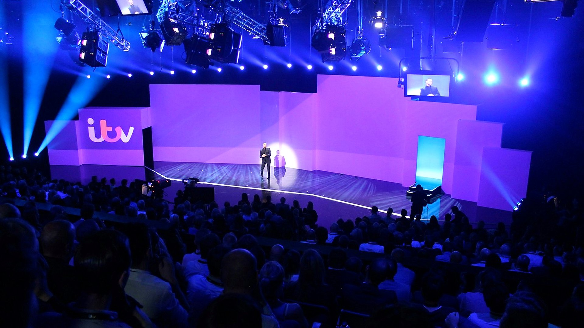 Wide view of the ITV Upfronts stage from the audience, with the ITV ident on stage left and a presenter centre stage.