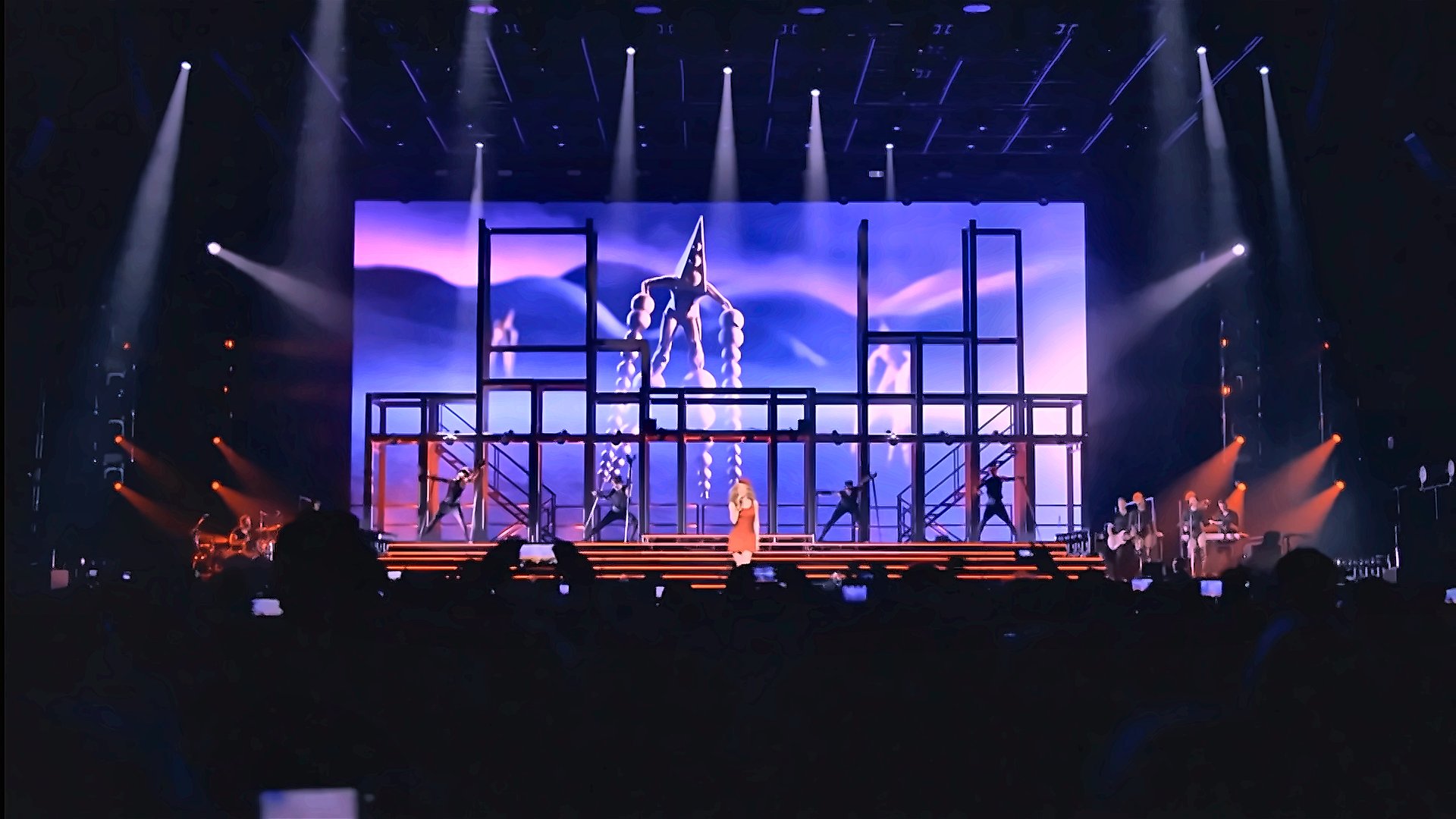Kylie Minogue performing in front of the staircase rig, with a pyramid-hatted dancer projected on the LED screen.