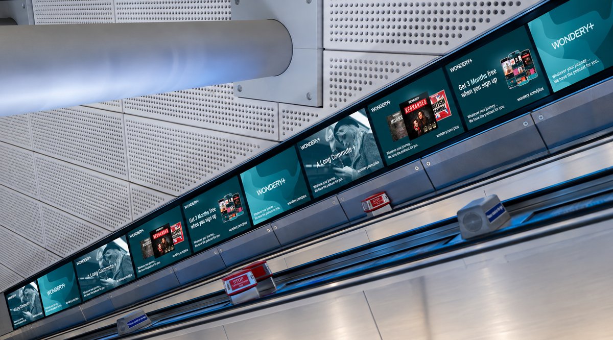 A low-angle view of a row of Wondery+ screens along an escalator bank, showing the app promotion and podcast artwork including Redhanded and British Scandal.